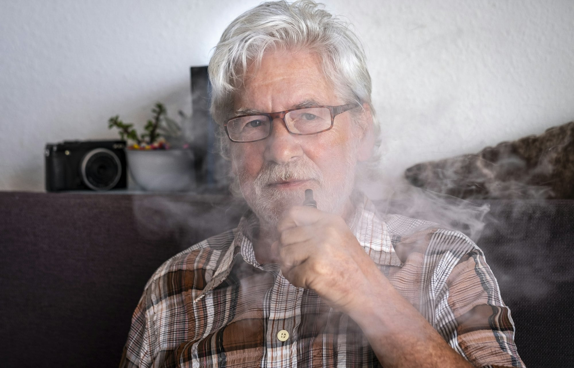 Bearded senior man smoking electronic cigarette surrounded by smoke, sitting on the sofa at home