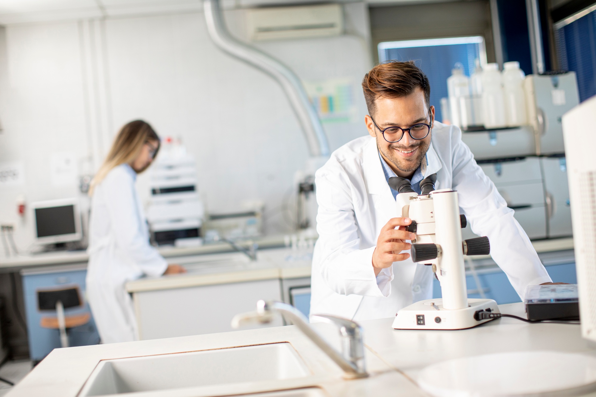 Young scientist in white lab coat working with binocular microscope in the material science lab